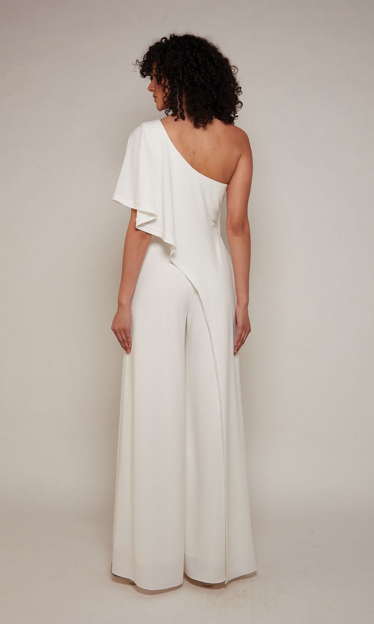 A person stands facing away, wearing the Alyce 70049 Long Formal Jumpsuit—a white, one-shoulder design with wide legs and an asymmetrical sleeve—creating an elegant cocktail look against a plain background.